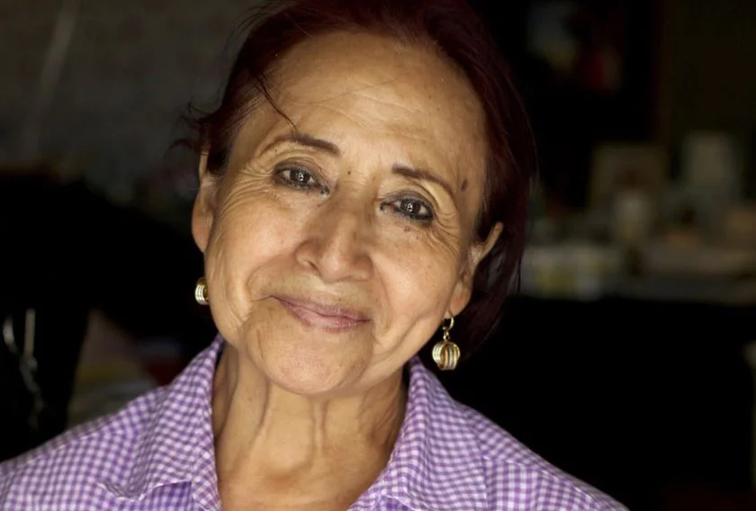 Hero - Vertical Image - Image Template Older woman with short hair and earrings smiling while wearing a purple checkered shirt.